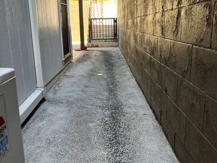Narrow walkway between buildings with concrete surface and brick wall, leading to a gate.
